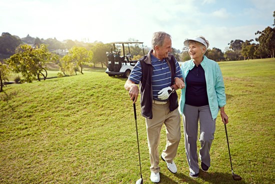 Smiling senior couple walking with arms locked  on gold course while carrying golf clubs