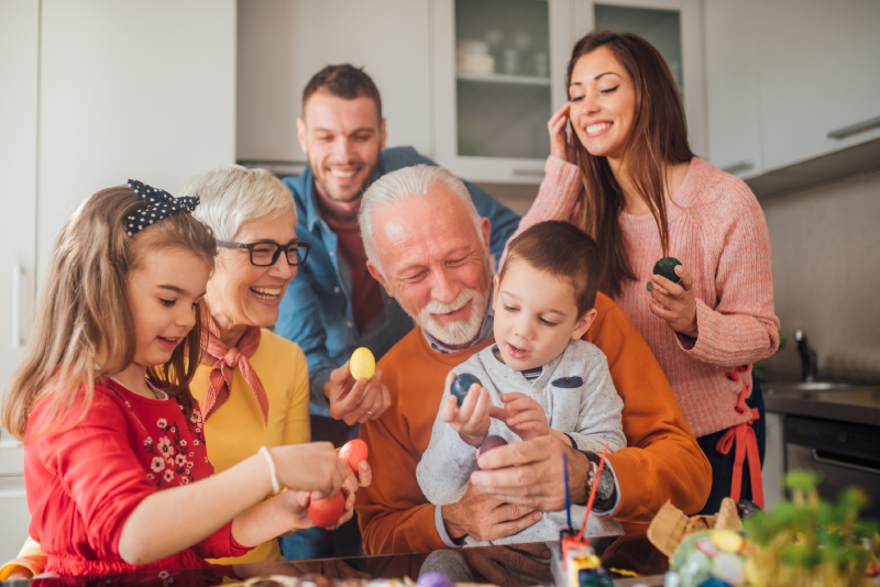 Senior couple holding their grandson and granddaughter on their laps as they paint Easter eggs