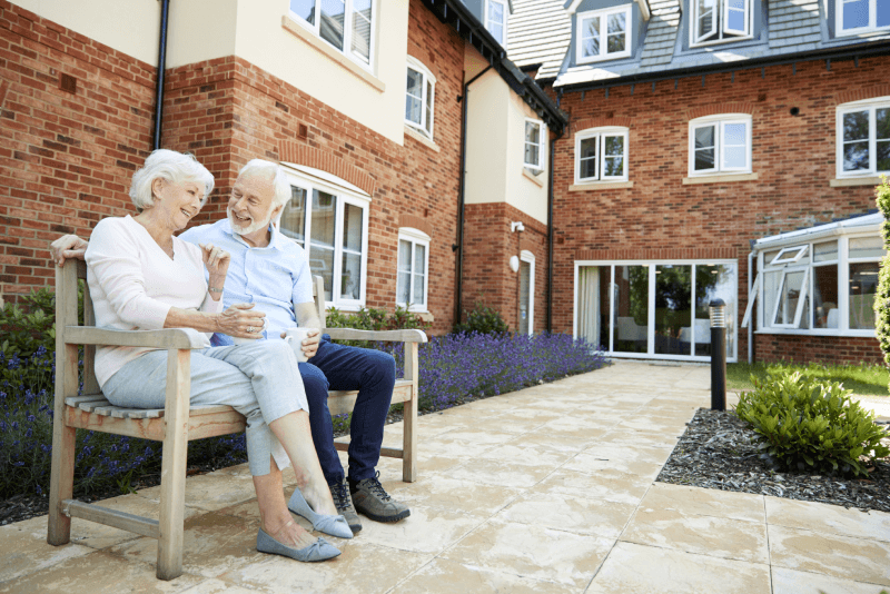 A happy senior couple sitting on a bench outside a continuing care retirement community.