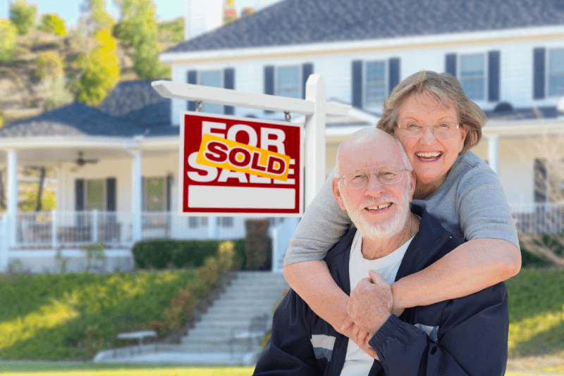 Senior Adult Couple in Front of Sold Home For Sale Real Estate Sign and Beautiful House.