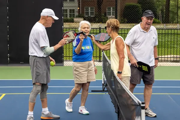 Four older adults stand on a pickleball court, two holding paddles by the net, appearing to chat or share a light moment during a game.