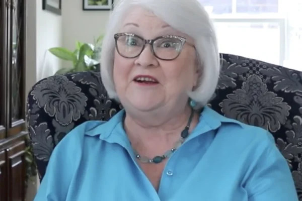 Older woman with white hair and glasses, wearing a light blue shirt and necklace, sits on a patterned chair in a well-lit room with plants and framed pictures in the background.