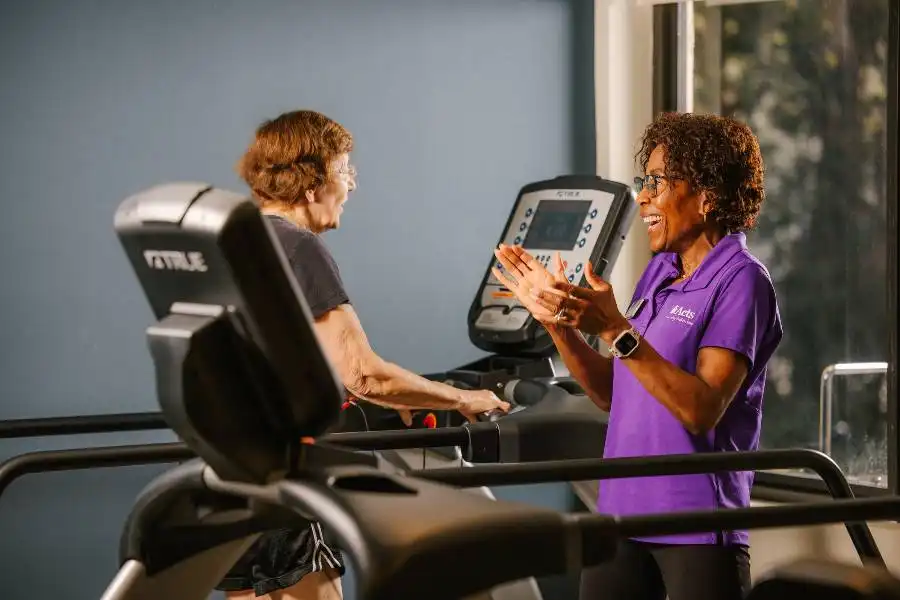 An older woman walks on a treadmill while a trainer in a purple shirt stands nearby, smiling and clapping in encouragement.