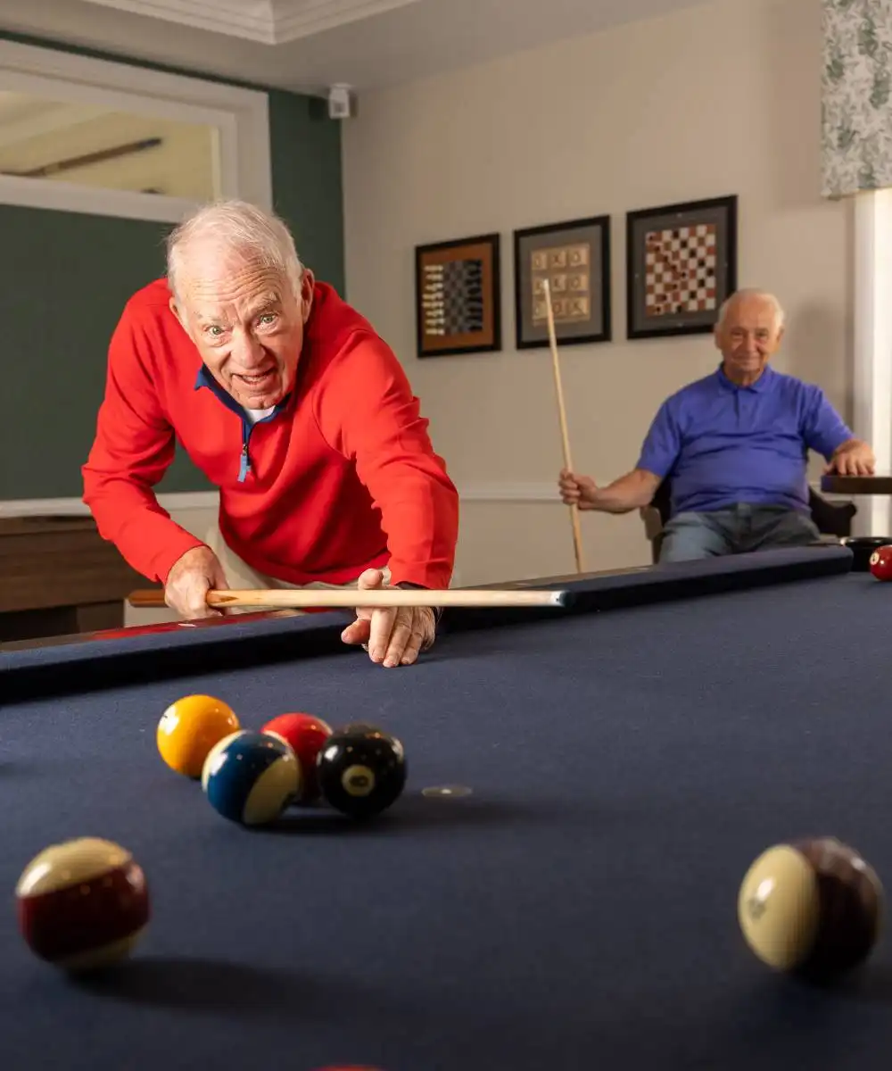 Two older men play pool in a room; one is about to take a shot while the other watches from a chair, holding a cue stick.