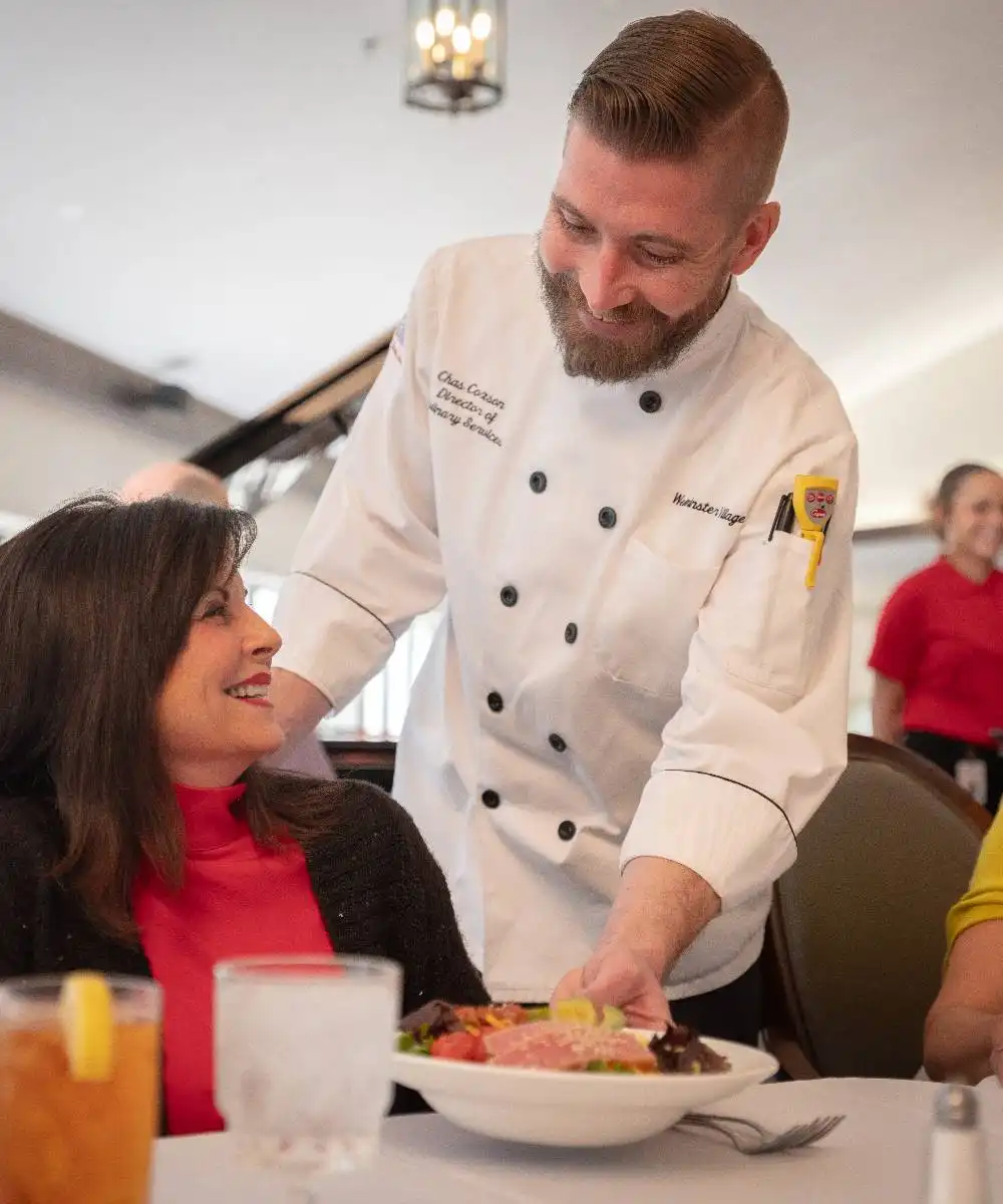 A chef in a white uniform serves a salad to a smiling woman seated at a restaurant table with drinks and another person nearby.
