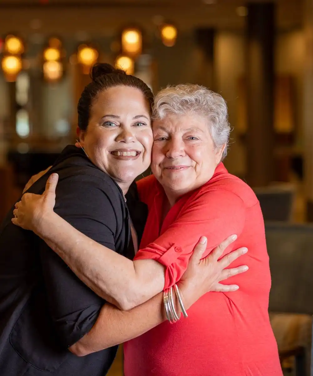 Two women, one younger and one older, smile and hug each other while standing indoors in a warmly lit room.