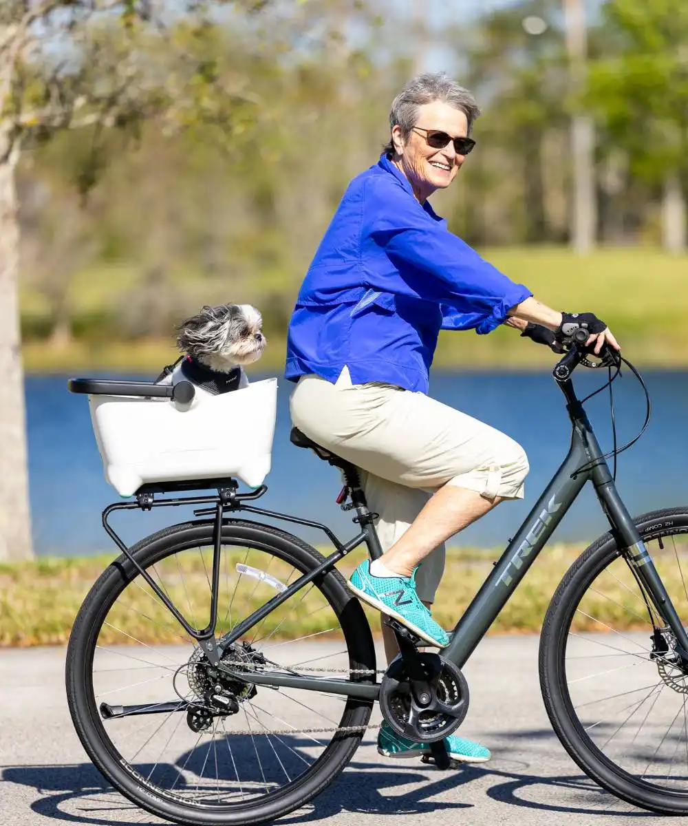An older woman rides a bicycle on a paved path with a small dog sitting in a white basket attached to the back.
