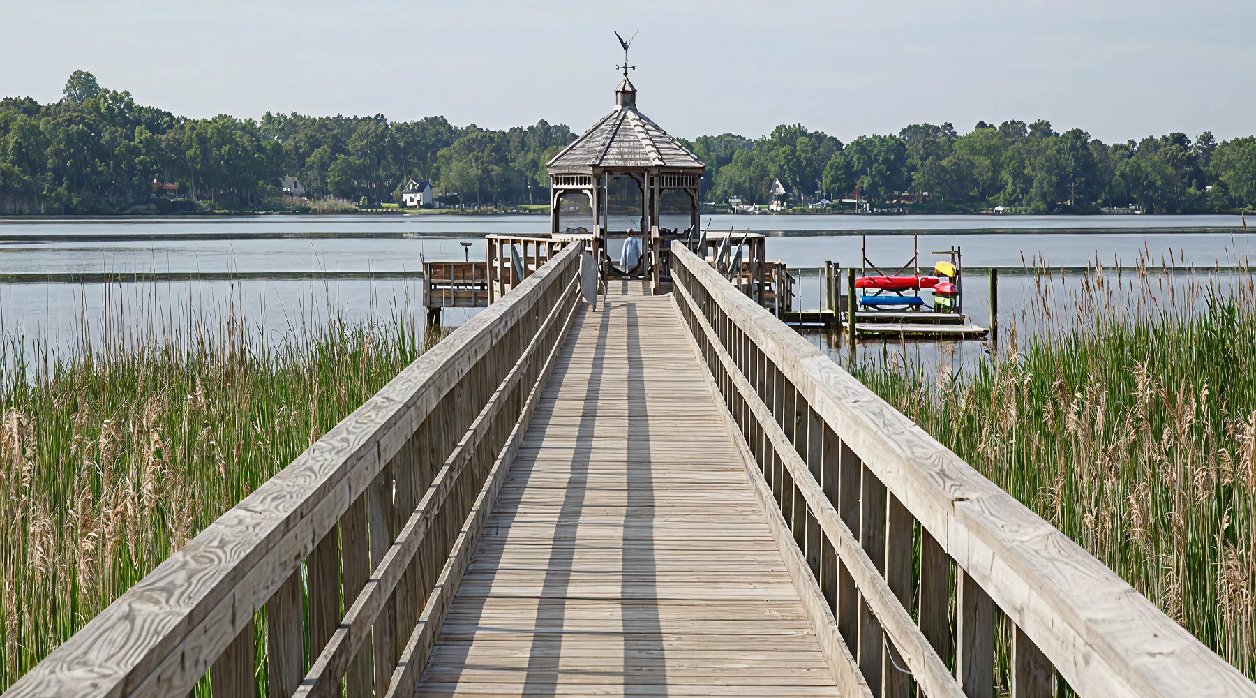 Wooden dock with railing leading to a gazebo over calm water, surrounded by reeds; kayaks and paddleboards are visible on a side platform.