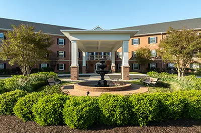 Three-story brick building with covered entrance, central fountain, and landscaped garden with benches in front.