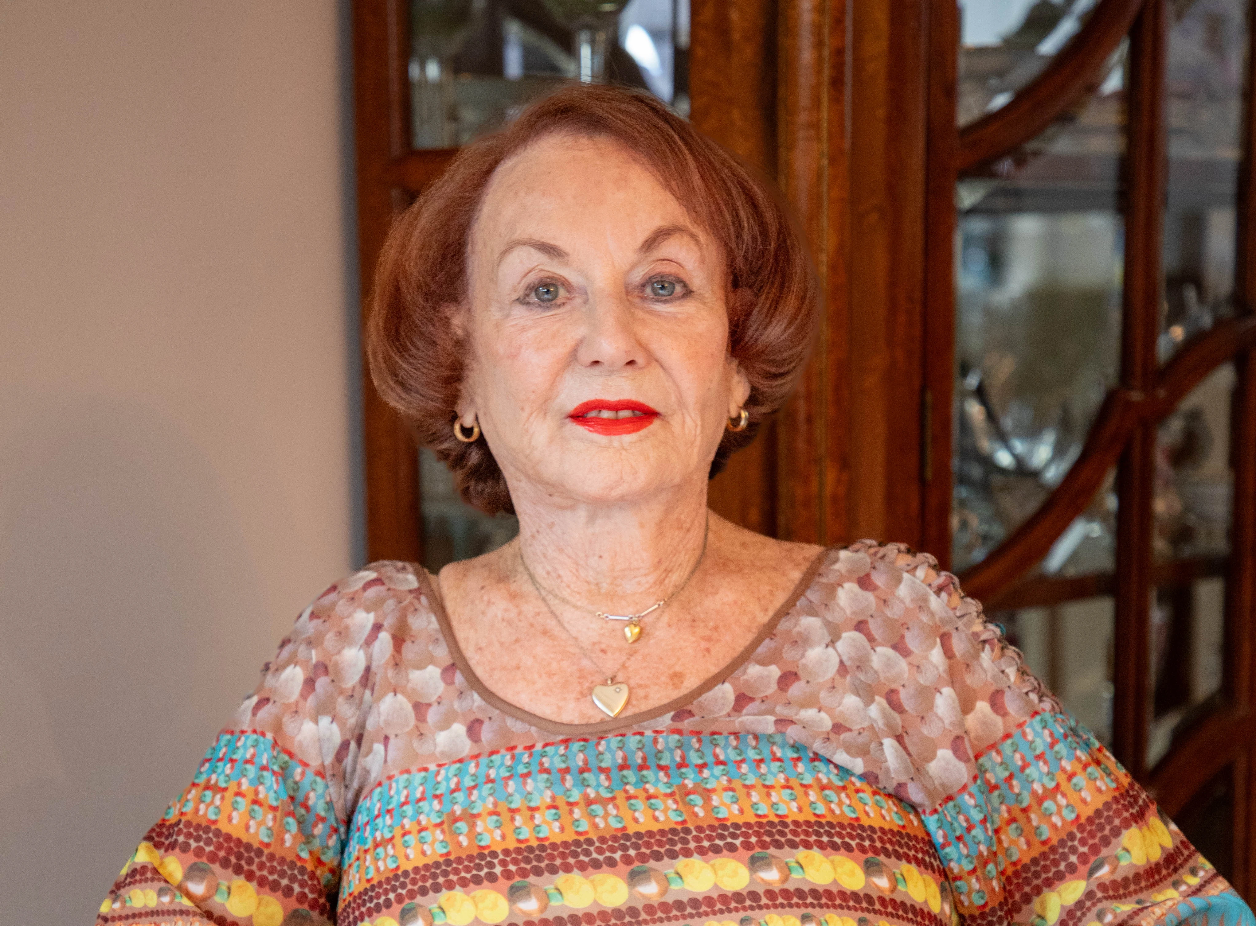An older woman with short red hair, wearing a colorful patterned top and gold jewelry, stands indoors in front of a wooden cabinet.
