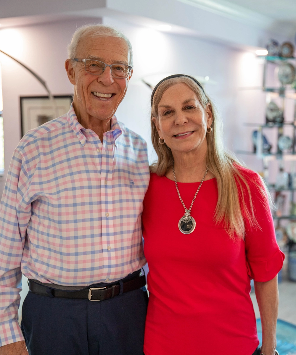 An older man in a plaid shirt and glasses stands next to a woman in a red top with long blonde hair and a pendant necklace, both smiling indoors.