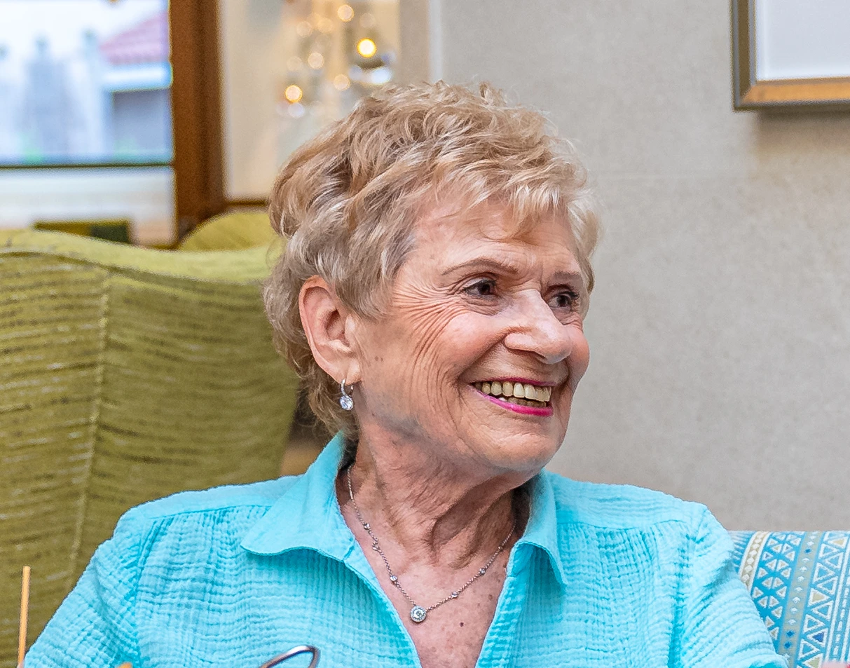 Older woman with short blonde hair, wearing a light blue shirt, sitting indoors and smiling to her right.