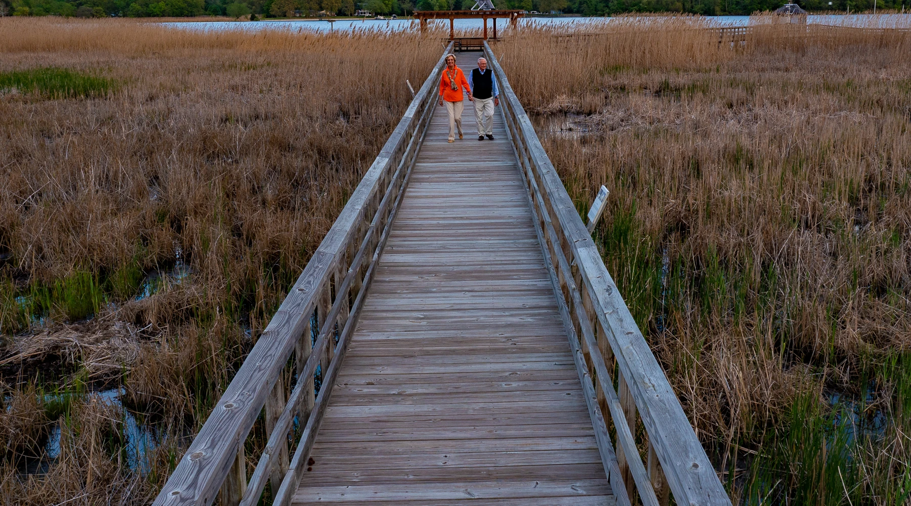 Two people walk side by side on a wooden boardwalk through tall grasses toward a covered dock over the water.