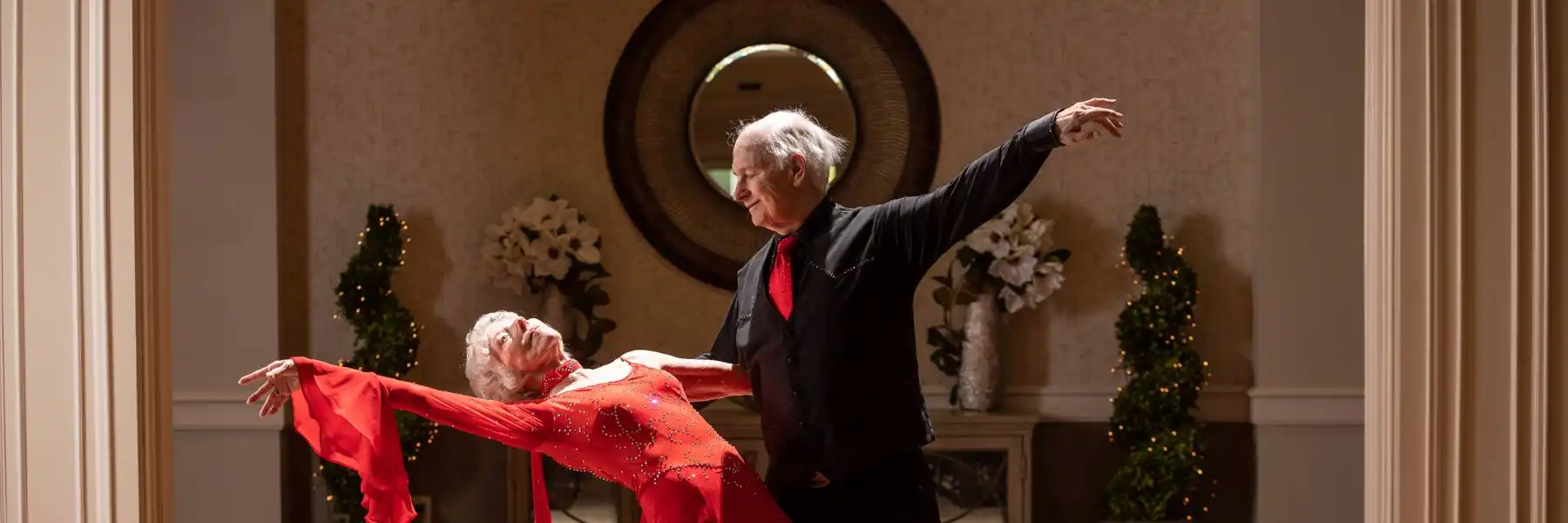 An elderly couple in formal attire perform a ballroom dance pose in a decorated room with a round mirror and flower arrangements in the background.