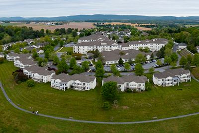 Aerial view of a large residential complex with multiple white buildings, green lawns, trees, and walking paths, set in a rural area with fields and hills in the background.