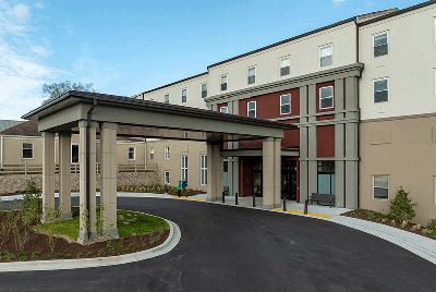 Three-story beige and red building with a covered entrance driveway, surrounded by landscaping, under a blue sky.