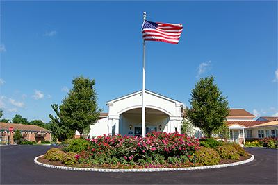 An American flag on a tall pole stands in front of a white building, surrounded by landscaped bushes and flowers under a clear blue sky.