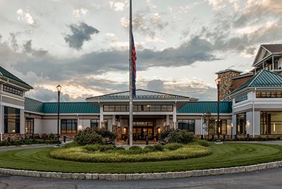 A large building with a green roof and a circular driveway is shown at dusk, with a flag at half-staff in front and cloudy sky overhead.