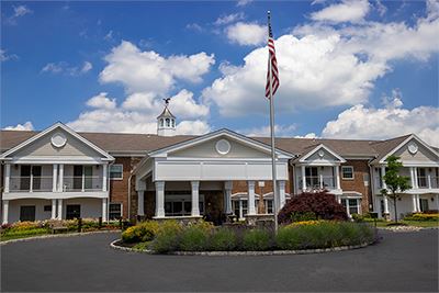 Two-story brick and white building with columns, an American flag on a pole in front, landscaped garden, and a partly cloudy sky in the background.