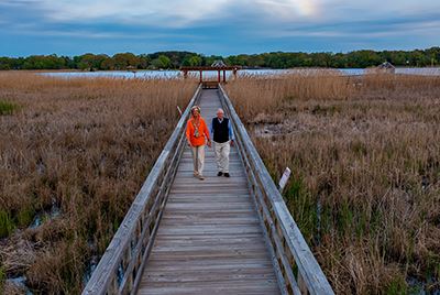 Two people walk on a wooden boardwalk through tall grass, heading toward a covered gazebo by a lake under a cloudy sky.