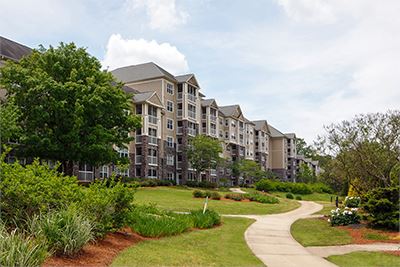 A wide sidewalk curves past landscaped grass and bushes in front of Gainesville senior apartments and carriage homes on a partly cloudy day.