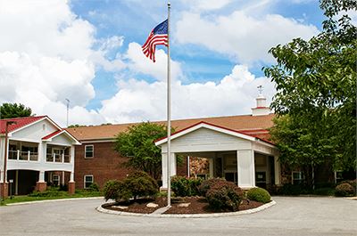 A brick building with a covered entrance and an American flag on a pole in front, surrounded by landscaped bushes under a partly cloudy sky.