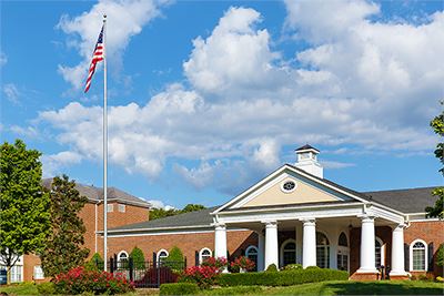 A brick building with white columns and a cupola, landscaped with bushes and flowers, and an American flag on a tall flagpole in front.
