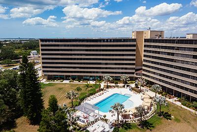 A large multi-story hotel with two wings surrounds a rectangular outdoor pool and palm trees, under a partly cloudy sky.