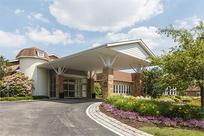 A building with a covered entrance, surrounded by landscaped flowers and trees, under a partly cloudy sky.