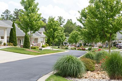 A residential street with single-story houses, green lawns, trees, and landscaped gardens along a paved road under a partly cloudy sky.