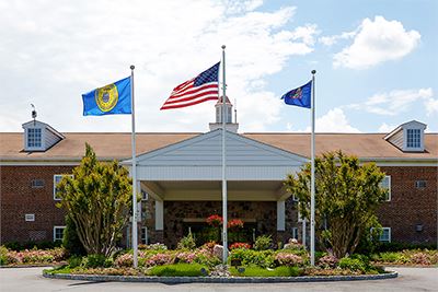 A brick building with three flagpoles out front displaying the American flag and two other flags, landscaped with bushes and flowers under a partly cloudy sky.