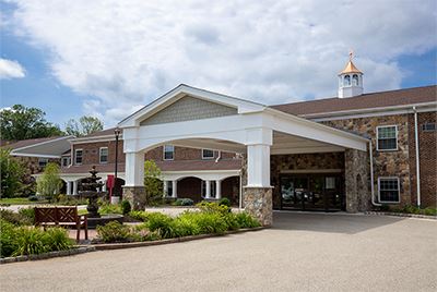 A large brick and stone building with a covered entrance, landscaped garden, and a small fountain in front, under a partly cloudy sky.
