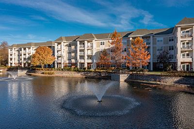 Three-story apartment building with balconies overlooks a pond with two fountains, surrounded by autumn trees under a blue sky.