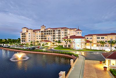 Large waterfront hotel with red-tiled roofs, several stories high, surrounded by a parking lot, palm trees, and a lit fountain in a pond at dusk.