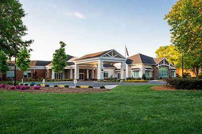 A large, single-story building with white pillars and a landscaped lawn, surrounded by trees under a clear sky.