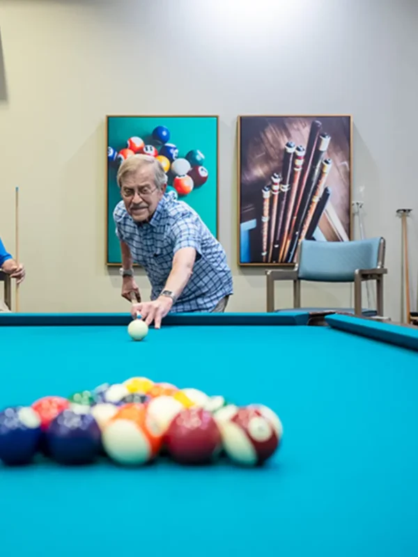 An older man in a blue checked shirt prepares to break on a billiards table, with colorful balls racked and framed pool-themed art on the wall.