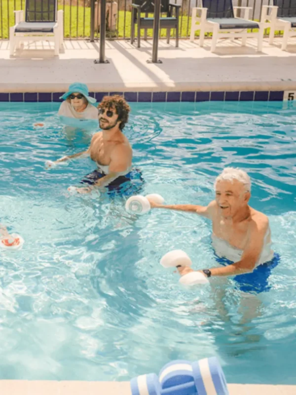 Three adults exercise with foam dumbbells in an outdoor pool. One person wears a sun hat and shirt, another wears goggles, and the third has gray hair and blue swim trunks.