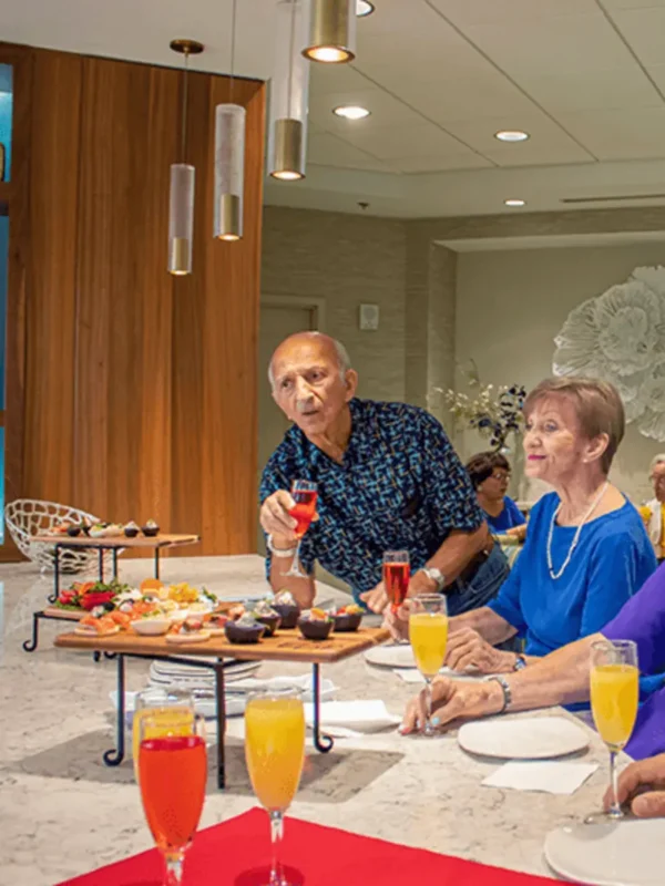A group of older adults sit around a table with assorted snacks and colorful drinks in a modern dining area.