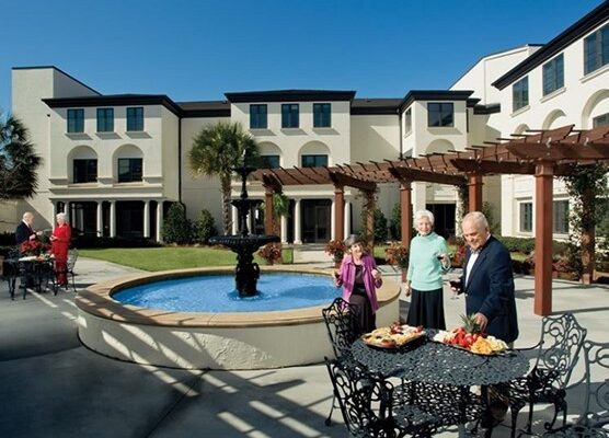 Several seniors stand by a round fountain and metal tables with food platters in a landscaped courtyard of a large residential building under a clear blue sky.