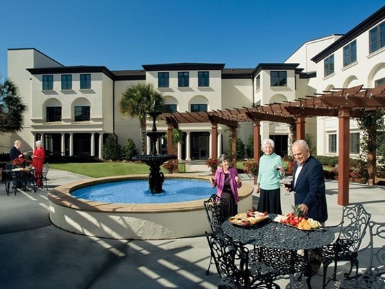 Several seniors stand by a round fountain and metal tables with food platters in a landscaped courtyard of a large residential building under a clear blue sky.