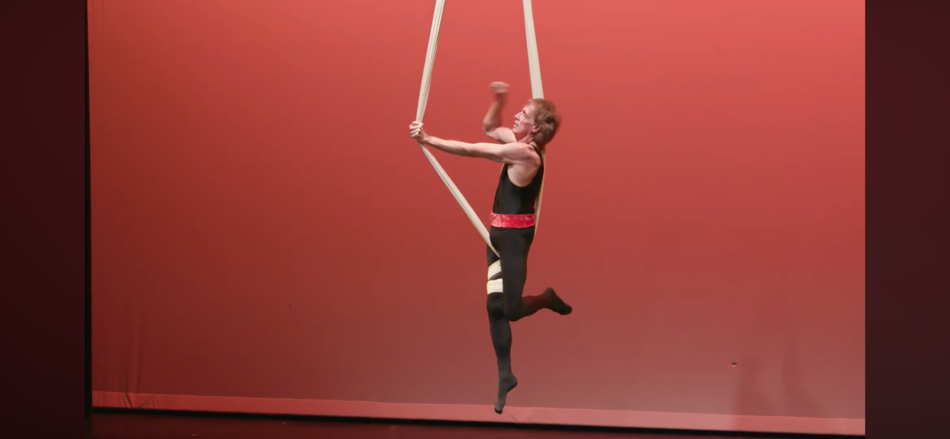A performer in a black and red costume hangs from aerial straps on a stage with a plain red background.
