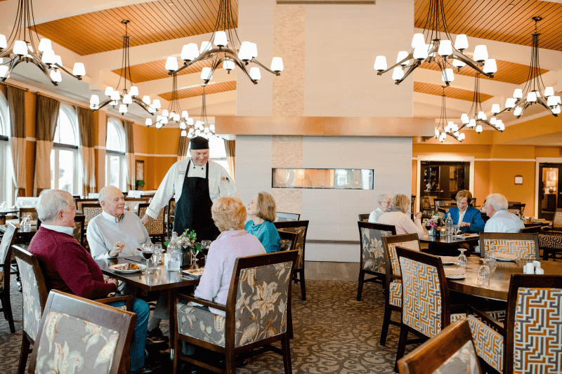 A group of seniors dine at a restaurant, engaged in conversation, while a chef stands by their table. Other diners are seated in the spacious, well-lit room.
