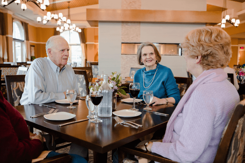 Three older adults sit at a restaurant table with drinks, talking and smiling. The table is set with plates, cutlery, and glasses. The setting appears to be upscale and well-lit.