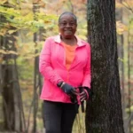 A woman in a pink jacket and gloves stands by a tree in a forest, holding walking poles. Leaves in the background show autumn colors.