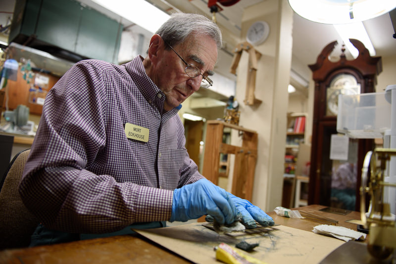 An older man wearing glasses, a name badge, and blue gloves works at a table, handling small tools and materials in a workshop with clocks and wooden shelves in the background.