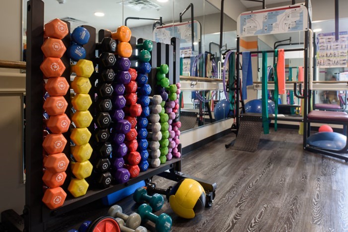 A rack of colorful dumbbells is arranged by weight in a gym with mirrors, exercise balls, resistance bands, and other workout equipment on a wooden floor.