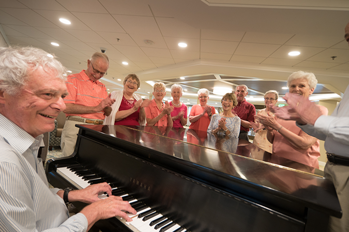 An older man plays the piano while a group of elderly people standing behind him smile and clap in a well-lit room.