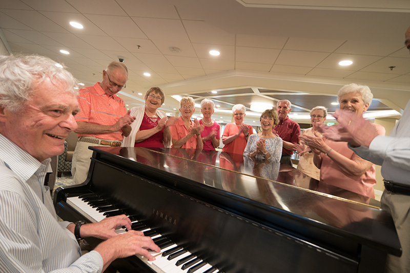 An elderly man plays piano while a group of smiling seniors stand around him, clapping and enjoying the performance in a well-lit room.