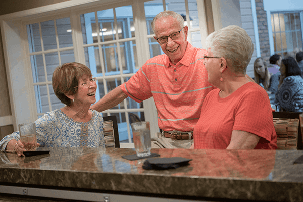 Three older adults sit at a kitchen counter, talking and smiling, with glasses of water in front of them. Other people are visible in the background through large windows.