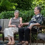 An older woman and man sit side by side on outdoor chairs, engaged in conversation, with greenery in the background.